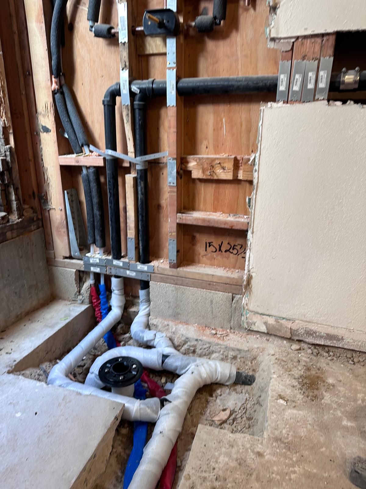 Bathroom rough-in with black drain and vent pipes in the open wall, a toilet flange in the floor and red and blue PEX water lines